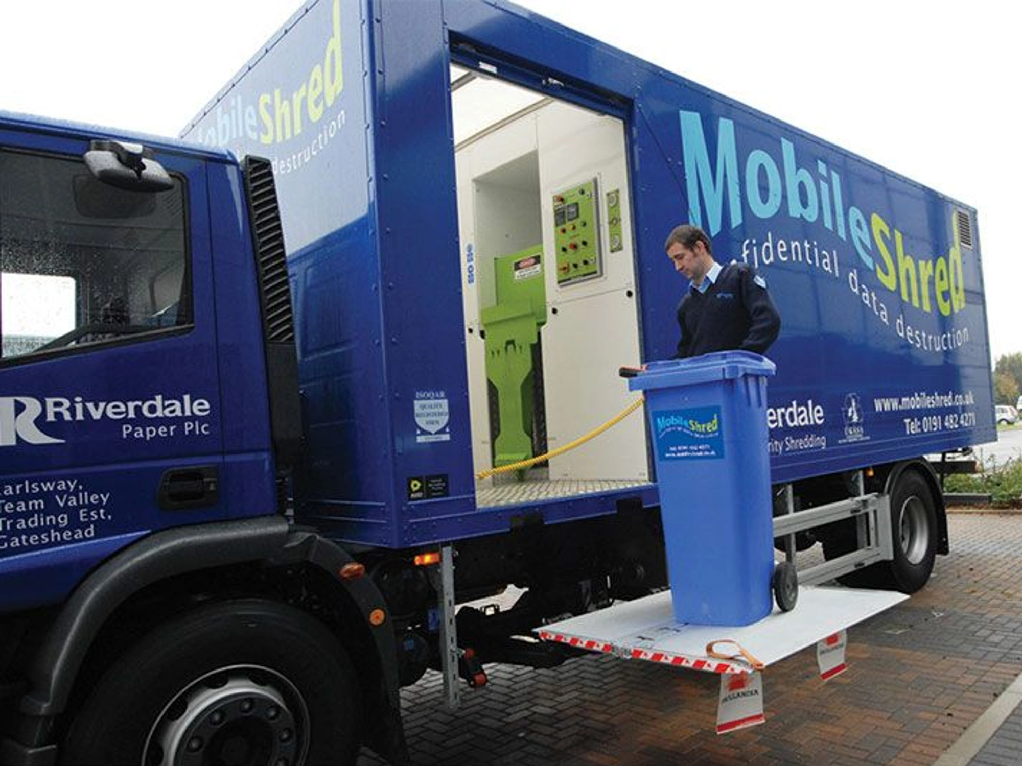 Blue mobile shredding van with Riverdale Recycling employee taking transporting wheelie bin onto van.