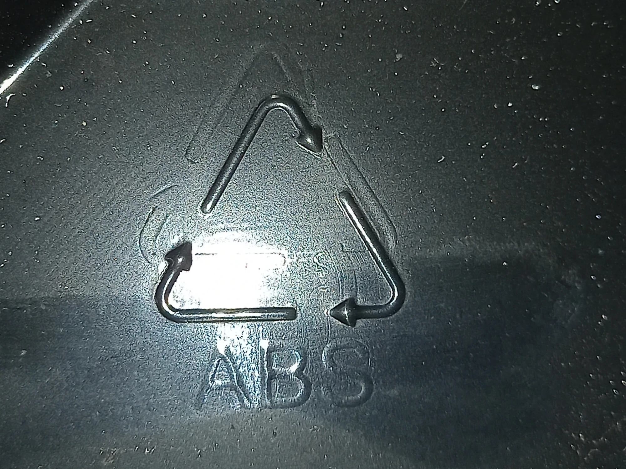 A textured, dark grey plastic surface shows a raised ABS recycling symbol.