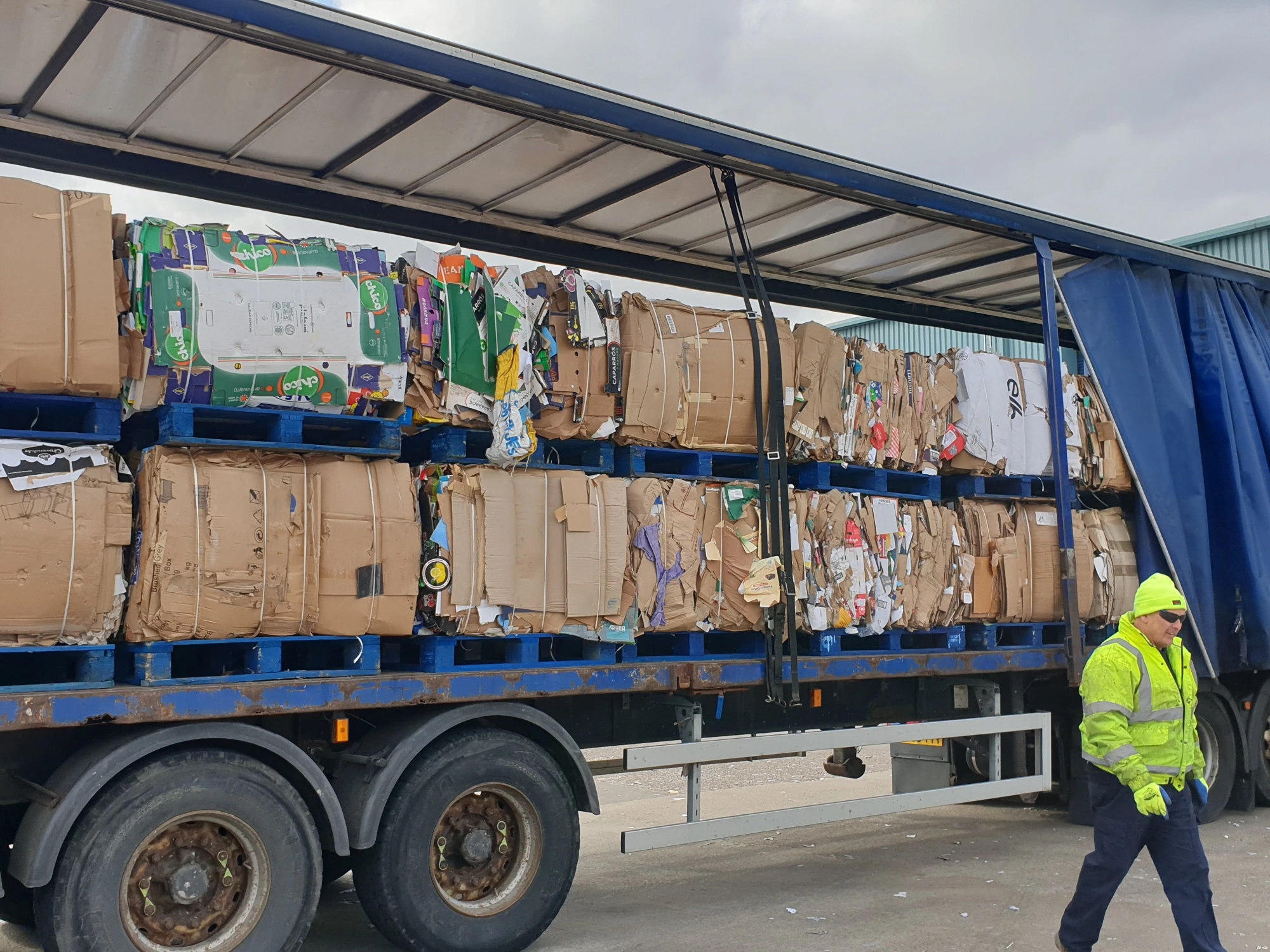A Riverdale Recycling truck with its side curtain pulled back, revealing large bales of compressed cardboard ready for transport.