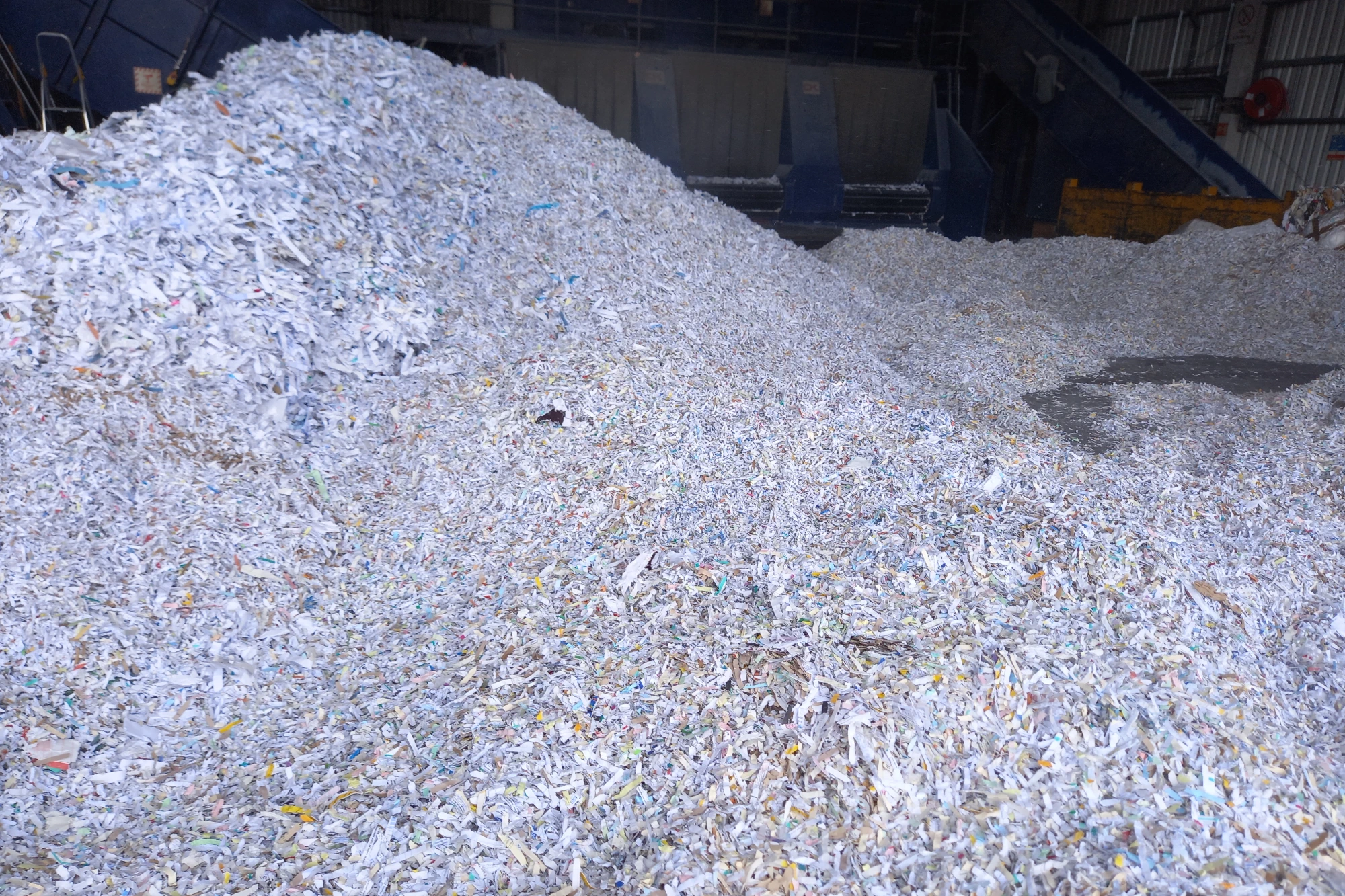 A pile of white and mixed-colour shredded paper fills the foreground of an industrial recycling facility.