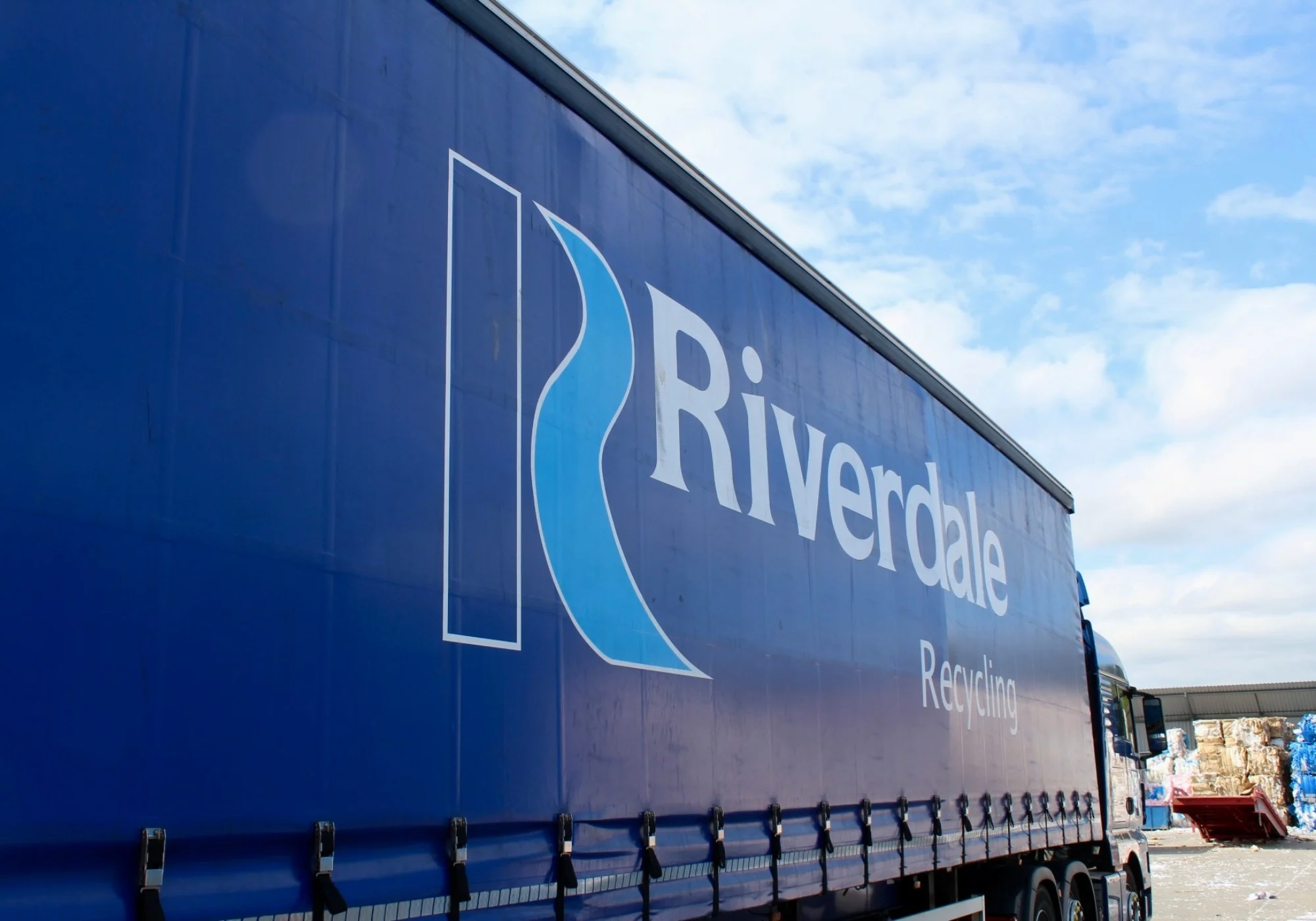 The side of a blue Riverdale Recycling curtain-side truck parked outdoors under a blue sky.