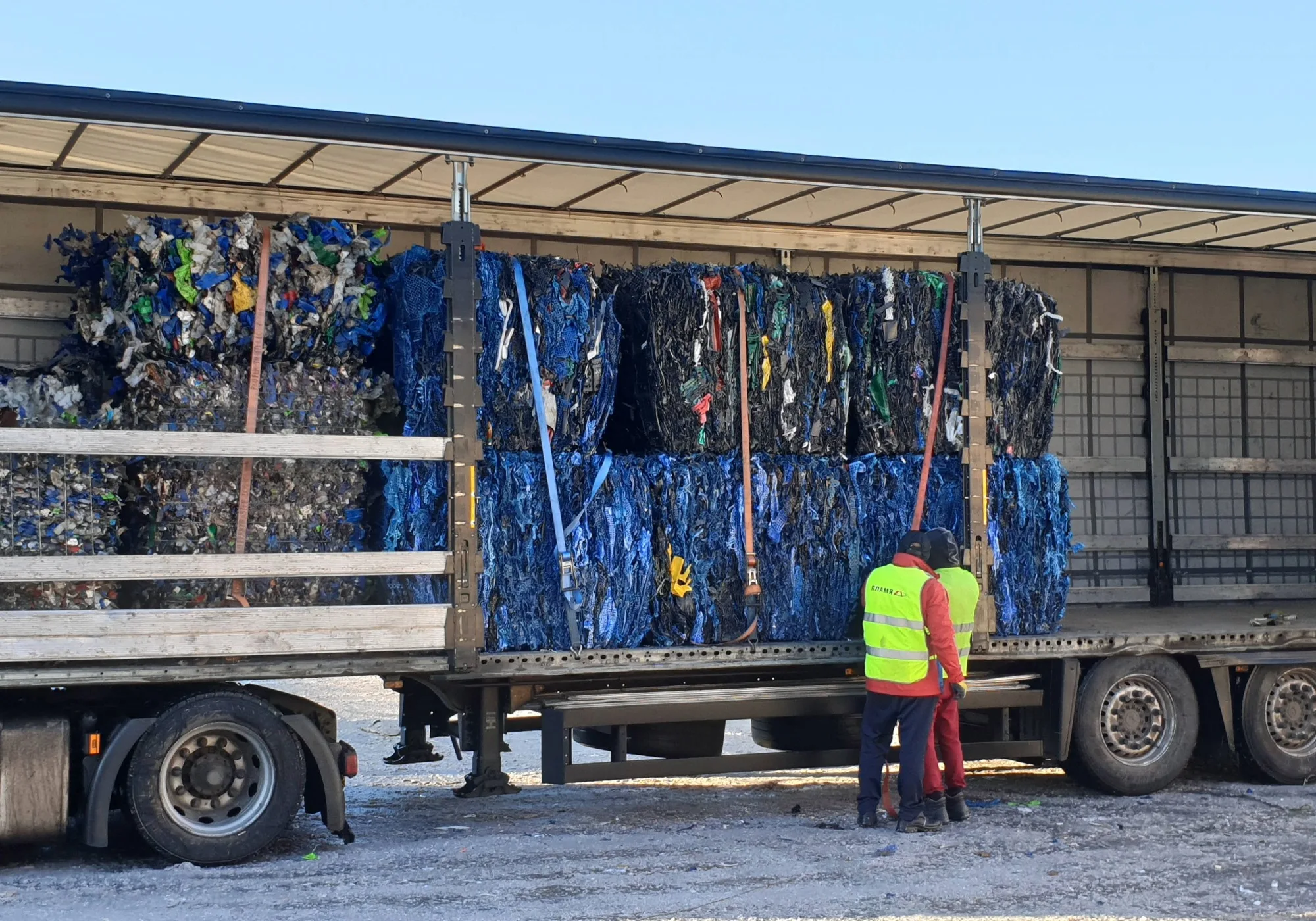 Two workers in high-visibility vests standing by a large truck loaded with stacked, strapped-down bales of blue and black recycled plastic.