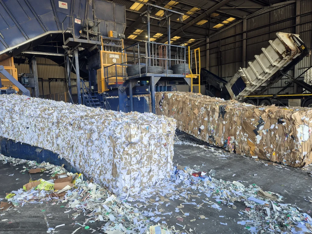 Inside a recycling facility, large bales of shredded paper and compacted cardboard sit in front of an industrial baler while a tipper lorry unloads recyclable materials for processing.