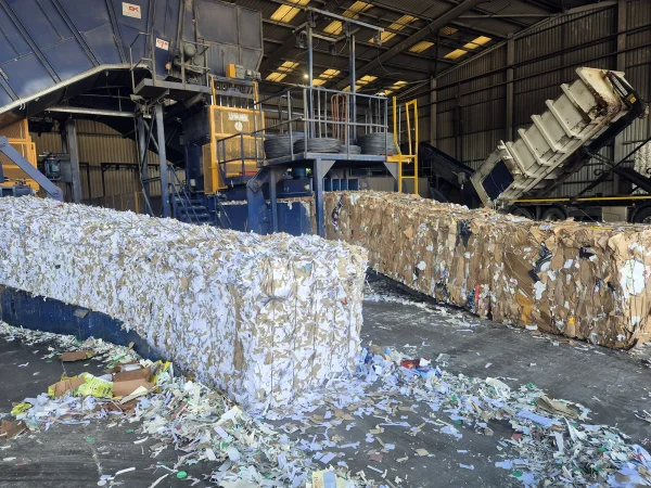 Inside a recycling facility, large bales of shredded paper and compacted cardboard sit in front of an industrial baler while a tipper lorry unloads recyclable materials for processing.